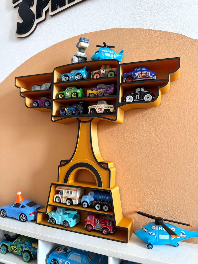 Puede incluir: Un estante de exhibici&oacute;n amarillo y negro con forma de trofeo, lleno de coches de juguete y un helic&oacute;ptero. El estante tiene m&uacute;ltiples compartimentos para guardar los veh&iacute;culos de juguete. El fondo es una pared de color naranja claro.