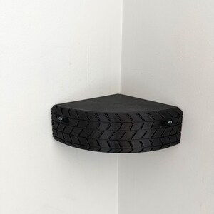 May include: A black, triangular corner shelf with a tire tread design. The shelf is mounted on a white wall and is secured with two silver screws. The shelf is designed to fit into a corner.