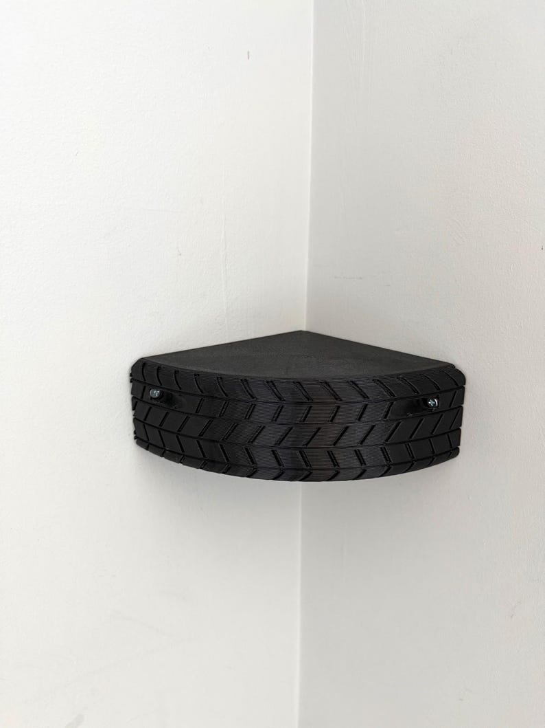 May include: A black, triangular corner shelf with a tire tread design. The shelf is mounted on a white wall and is secured with two silver screws. The shelf is designed to fit into a corner.