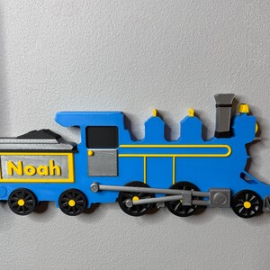 3D-printed blue train engine with yellow and silver details, labelled “Noah” on the coal car. Displayed next to a toy car shelf to give the impression of pulling it like a train.