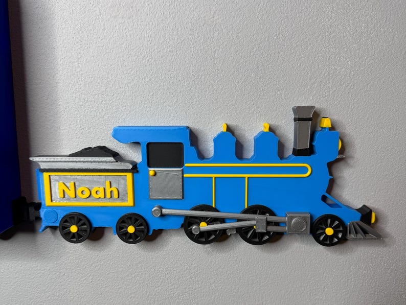 3D-printed blue train engine with yellow and silver details, labelled “Noah” on the coal car. Displayed next to a toy car shelf to give the impression of pulling it like a train.