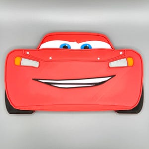 K&ouml;nnte beinhalten: Ein rotes, Lightning McQueen-f&ouml;rmiges Objekt mit einem l&auml;chelnden Mund, blauen Augen und wei&szlig;en Scheinwerfern. Das Auto hat schwarze Reifen und eine wei&szlig;e Umrandung um den Mund. Bei dem Artikel handelt es sich wahrscheinlich um ein Spielzeug oder ein dekoratives St&uuml;ck.