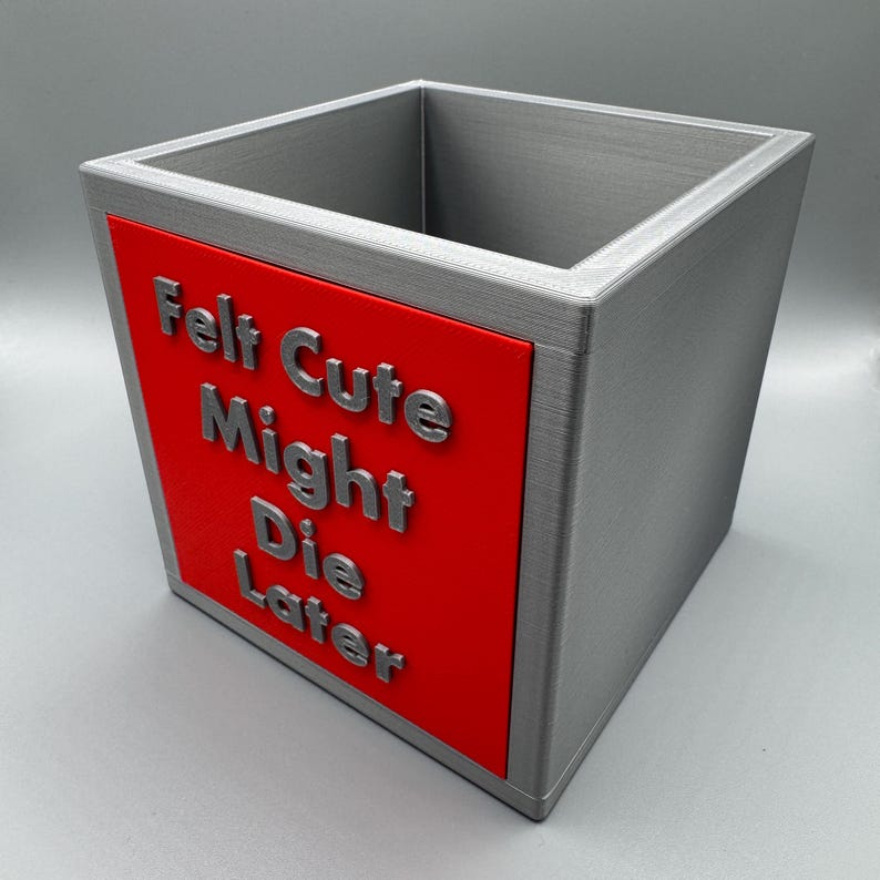 May include: A square, gray container with a red panel. The red panel features the phrase "Felt Cute Might Die Later" in raised, silver lettering. The container has a textured appearance.