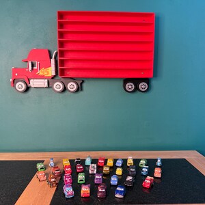 May include: A red wall-mounted display shelf shaped like a semi-truck with a trailer featuring multiple horizontal slots for small toy cars. The truck cab has a yellow number 95 graphic. Below the shelf, a collection of small toy cars is arranged.