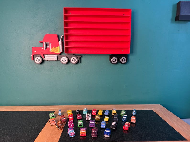 May include: A red wall-mounted display shelf shaped like a semi-truck with a trailer featuring multiple horizontal slots for small toy cars. The truck cab has a yellow number 95 graphic. Below the shelf, a collection of small toy cars is arranged.