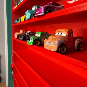 May include: A bright red wall-mounted display shelf shaped like a truck, featuring multiple horizontal tiers. Several small, colorful toy cars with cartoon-style eyes are arranged on the shelves, including orange, green, purple, and blue vehicles.