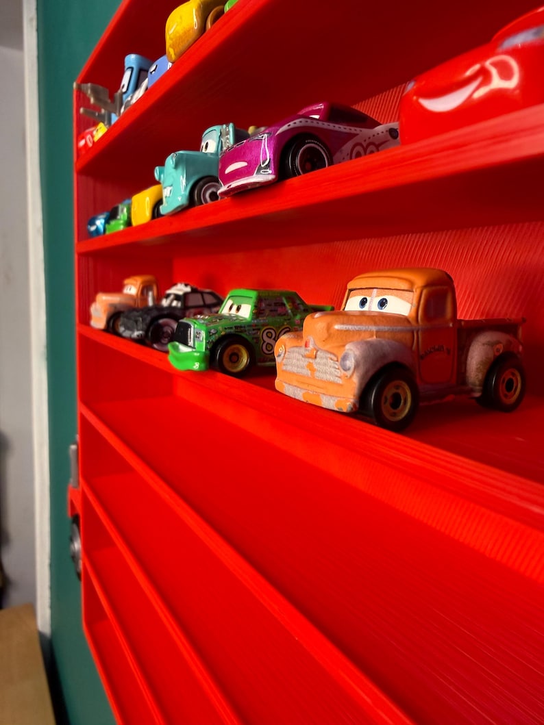 May include: A bright red wall-mounted display shelf shaped like a truck, featuring multiple horizontal tiers. Several small, colorful toy cars with cartoon-style eyes are arranged on the shelves, including orange, green, purple, and blue vehicles.