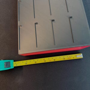 May include: A black jewelry display tray with multiple slots, a red base, and a yellow measuring tape. The tray appears to be made of plastic or a similar material. The tape measure shows measurements in inches.