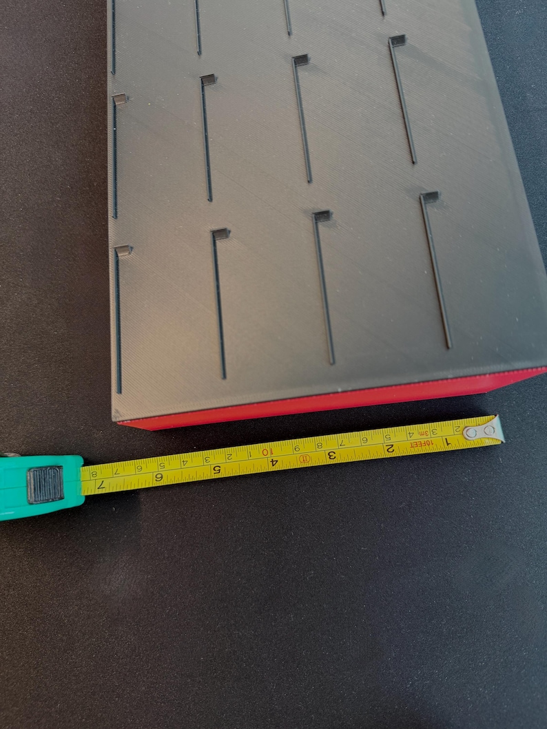 May include: A black jewelry display tray with multiple slots, a red base, and a yellow measuring tape. The tray appears to be made of plastic or a similar material. The tape measure shows measurements in inches.