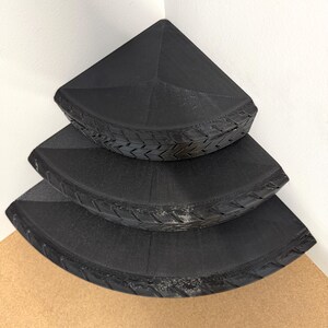 May include: Three black corner shelves stacked on top of each other. The shelves are triangular with a textured, tire-like design around the edges. The shelves are placed in a corner against a white wall.
