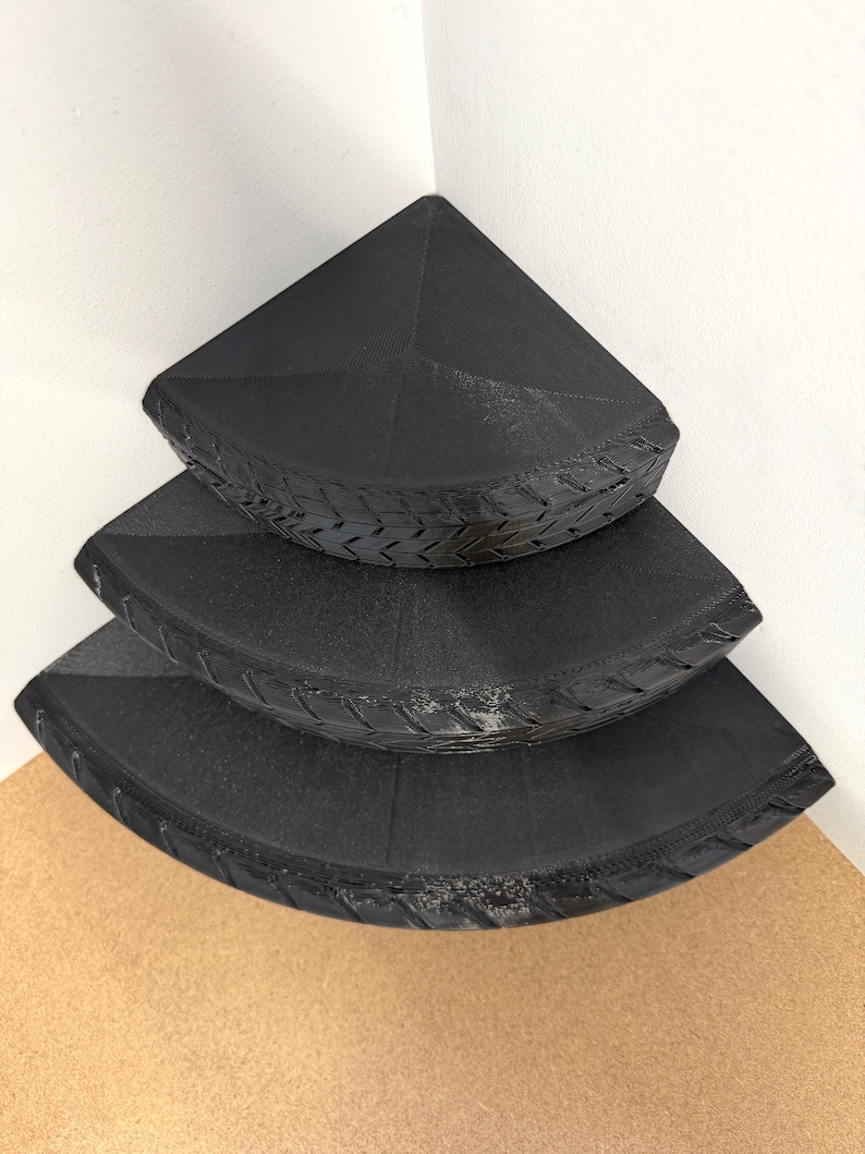May include: Three black corner shelves stacked on top of each other. The shelves are triangular with a textured, tire-like design around the edges. The shelves are placed in a corner against a white wall.