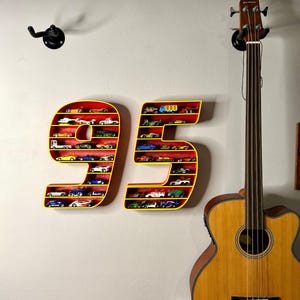 May include: A wall-mounted display shelf shaped like the number 95, painted yellow with a red interior. The shelves are filled with miniature toy cars of various colors. A guitar is in the background.