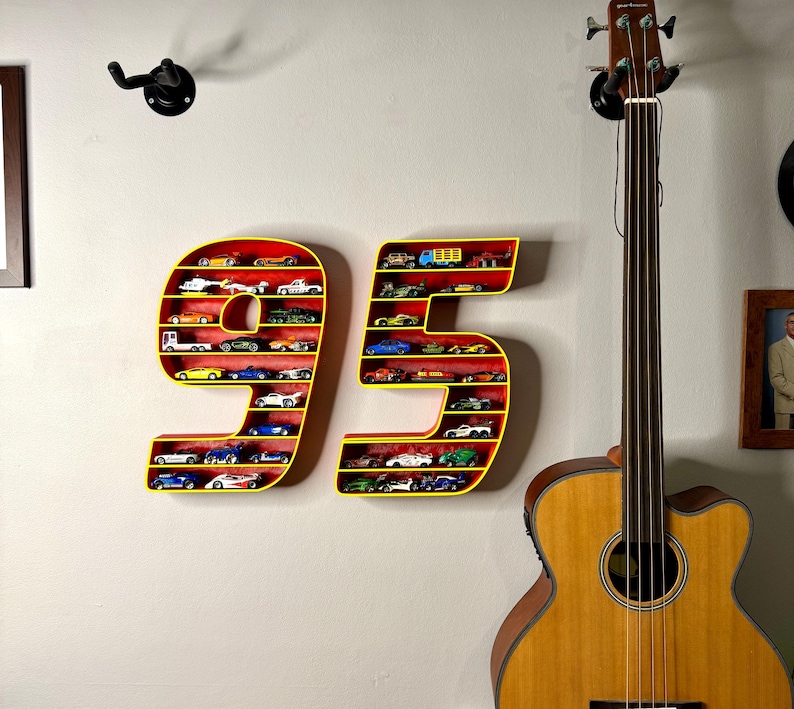 May include: A wall-mounted display shelf shaped like the number 95, painted yellow with a red interior. The shelves are filled with miniature toy cars of various colors. A guitar is in the background.