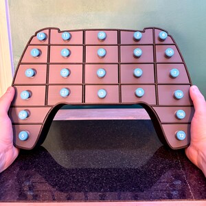May include: A brown advent calendar in the shape of a video game controller. It has 24 small drawers with blue knobs, each numbered from 1 to 24. The controller is held by two hands.