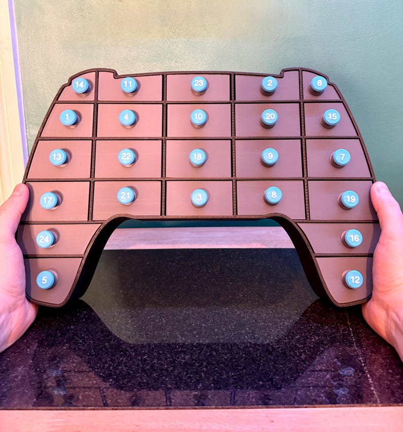 May include: A brown advent calendar in the shape of a video game controller. It has 24 small drawers with blue knobs, each numbered from 1 to 24. The controller is held by two hands.