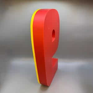 May include: A large, red letter "P" with a yellow edge, standing upright. The letter has a rounded shape and a hole in the center. The background is a solid, light gray.