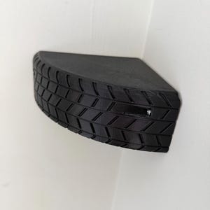 May include: A black, corner-mounted shelf with a tire tread design. The shelf is a quarter-circle shape and appears to be made of a solid material. The tire tread pattern is visible on the front edge, adding a unique texture to the shelf.