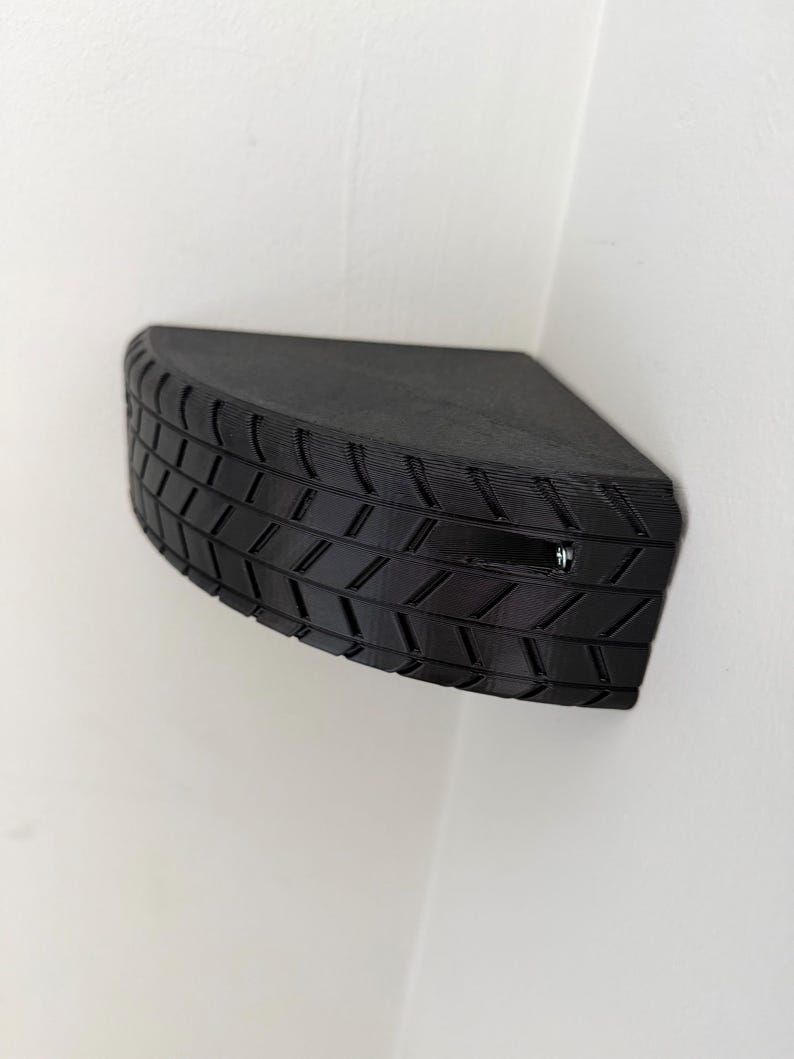 May include: A black, corner-mounted shelf with a tire tread design. The shelf is a quarter-circle shape and appears to be made of a solid material. The tire tread pattern is visible on the front edge, adding a unique texture to the shelf.