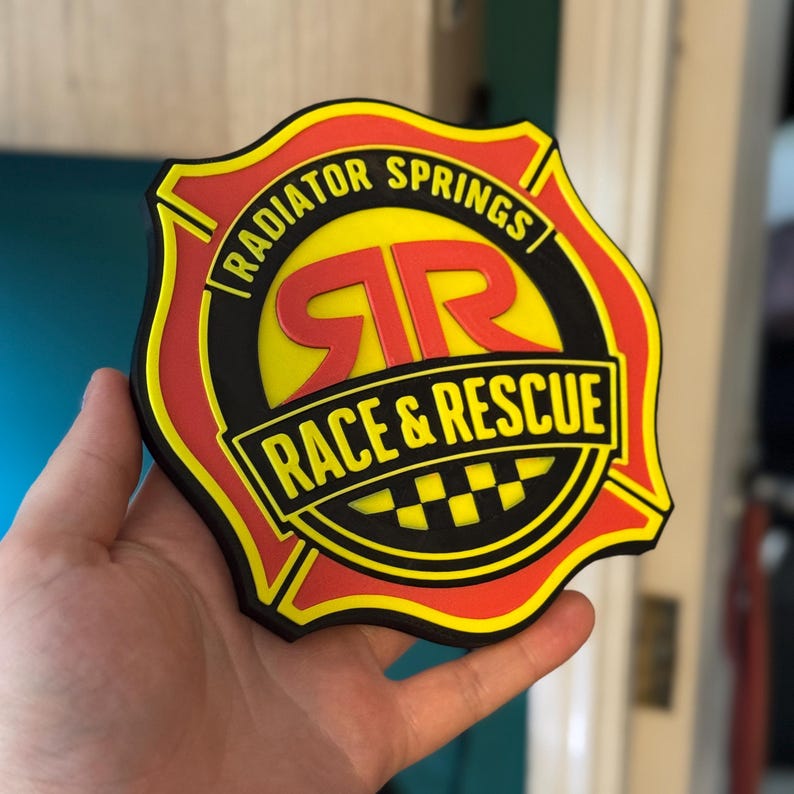 May include: A colorful, die-cut emblem featuring the text "RADIATOR SPRINGS RACE & RESCUE." The design includes a yellow center with red lettering and a black border. The emblem is shaped like a badge.