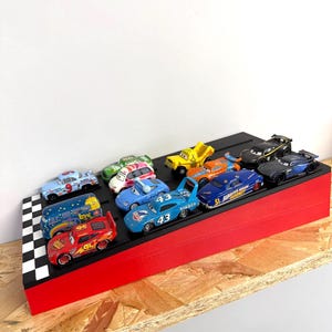 May include: A collection of toy race cars in various colors, including red, blue, and yellow, are displayed on a black and red wooden shelf. The shelf has a checkered flag design on one end. The cars are arranged in rows.