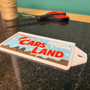 K&ouml;nnte beinhalten: Ein rechteckiges wei&szlig;es Schild mit den Worten "Welcome to Cars Land" in Rot und Blau, mit einem Weihnachtsmannhut auf dem Buchstaben "C". Das Schild tr&auml;gt auch die Worte "Season's Greetings".