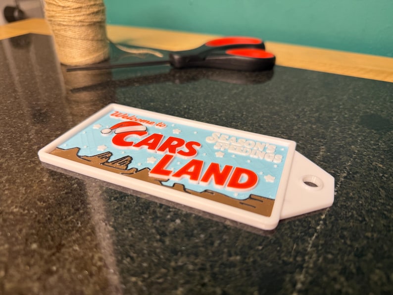 K&ouml;nnte beinhalten: Ein rechteckiges wei&szlig;es Schild mit den Worten "Welcome to Cars Land" in Rot und Blau, mit einem Weihnachtsmannhut auf dem Buchstaben "C". Das Schild tr&auml;gt auch die Worte "Season's Greetings".