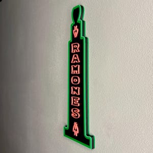 Puede incluir: Un letrero negro y verde con la palabra "RAMONES" en letras rosas. El letrero tiene una forma &uacute;nica con un contorno verde y est&aacute; montado en una pared gris claro. El letrero es un objeto decorativo.