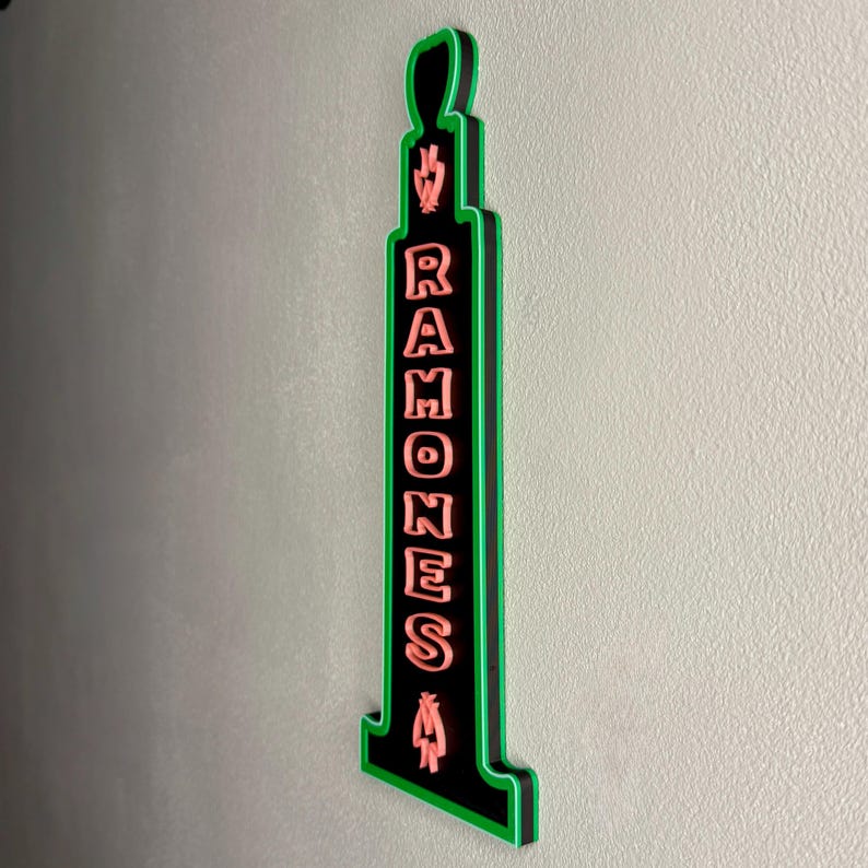 Puede incluir: Un letrero negro y verde con la palabra "RAMONES" en letras rosas. El letrero tiene una forma &uacute;nica con un contorno verde y est&aacute; montado en una pared gris claro. El letrero es un objeto decorativo.