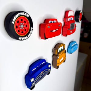 May include: A collection of colorful car-themed magnets, including a black tire with "LIGHTYEAR" text, a red race car, a red truck, and other car designs in blue, orange, and brown. These magnets are likely for a refrigerator or magnetic surface.