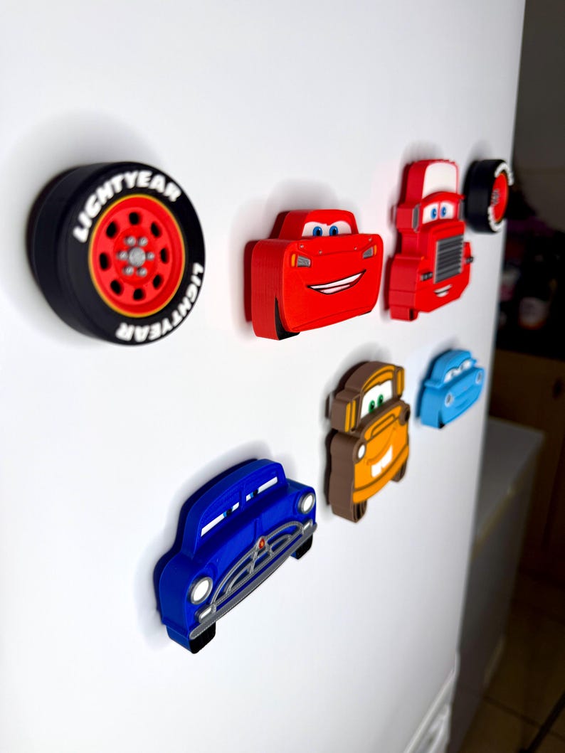 May include: A collection of colorful car-themed magnets, including a black tire with "LIGHTYEAR" text, a red race car, a red truck, and other car designs in blue, orange, and brown. These magnets are likely for a refrigerator or magnetic surface.