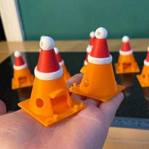 Może przedstawiać: Małe pomarańczowe pachołki drogowe, każdy zwieńczony czerwoną czapką Świętego Mikołaja i białym wykończeniem. Stożki mają kwadratową podstawę i mały otw&oacute;r z boku. Każdy kapelusz jest zwieńczony białym pomponem. Widocznych jest kilka stożk&oacute;w.