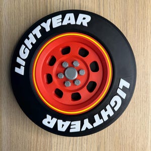 May include: A black and red decorative wheel with the text "LIGHTYEAR" in white. The wheel has a red center with silver bolts and a yellow stripe around the rim. The design is reminiscent of a race car wheel.