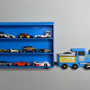 3D-printed toy train engine labelled “Noah” placed next to a matching blue toy car shelf, giving the illusion the train is pulling it. The shelf is empty and mounted on a grey wall.