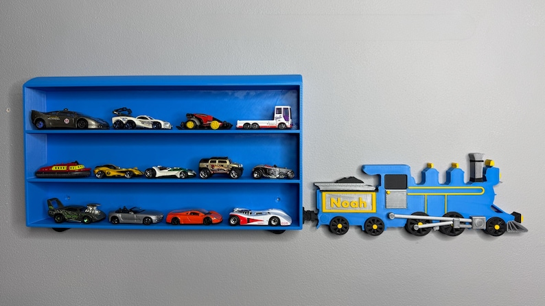 3D-printed toy train engine labelled “Noah” placed next to a matching blue toy car shelf, giving the illusion the train is pulling it. The shelf is empty and mounted on a grey wall.