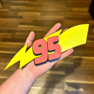 May include: A red and black number 95, with a black outline, is set against a yellow lightning bolt shape. The design is held in a hand, with a wooden background.