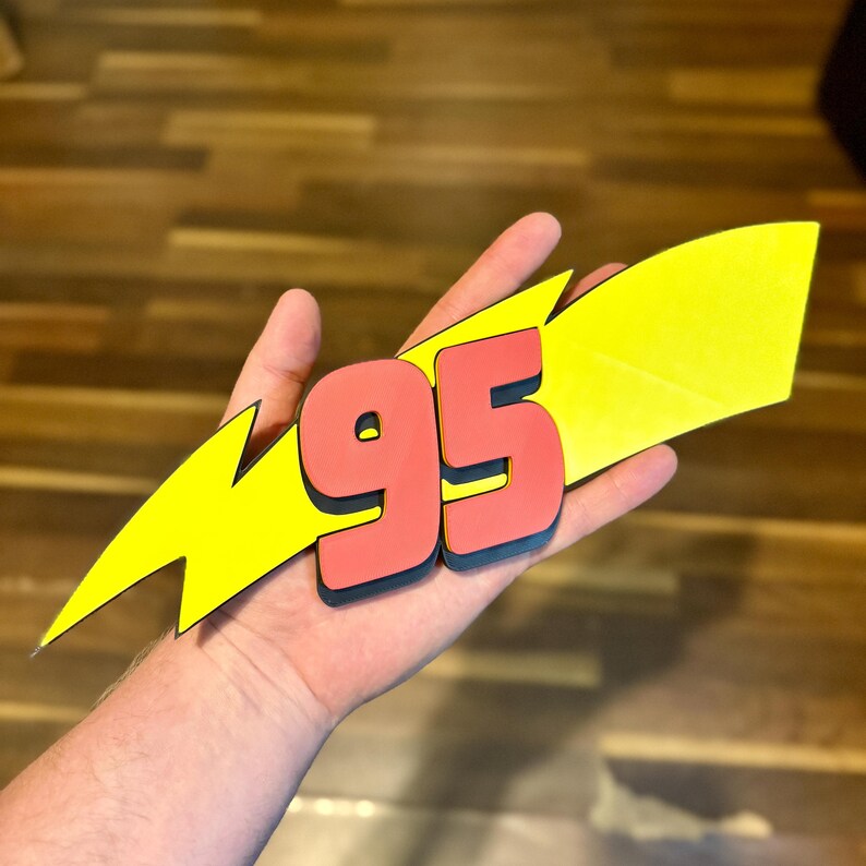 May include: A red and black number 95, with a black outline, is set against a yellow lightning bolt shape. The design is held in a hand, with a wooden background.