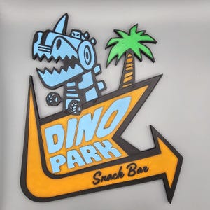 May include: A Dino Park sign featuring a cartoon dinosaur and palm tree. The sign is an orange arrow shape with black trim and the words "Dino Park" in blue. The words "Snack Bar" are below.