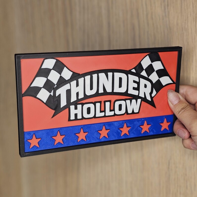 May include: Rectangular sign featuring the words "THUNDER HOLLOW" in white, above a blue stripe with orange stars. The sign has a red background and two chequered flags. The sign is held by a hand.