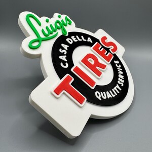 May include: A 3D-printed sign with the text "Luigis" in green script above a black circle with the words "Casa Della Tires Quality Service" and red "TIRES" lettering. The sign is white with a gray background.