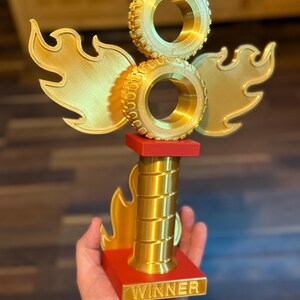 May include: A gold trophy featuring two stacked tyre-shaped elements, flame-like wings, and a red base. The base has the word "WINNER" in gold lettering. The trophy is held in a hand.