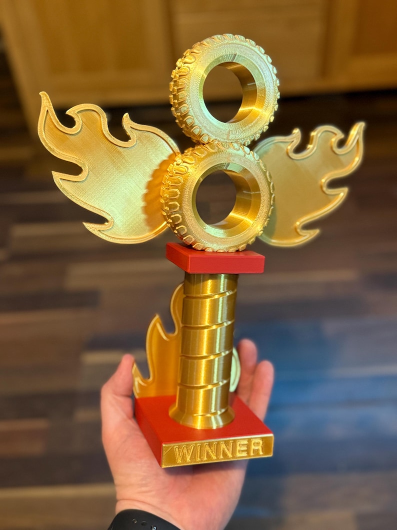 May include: A gold trophy featuring two stacked tyre-shaped elements, flame-like wings, and a red base. The base has the word "WINNER" in gold lettering. The trophy is held in a hand.