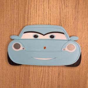 May include: A light blue cartoon car with a smiling face, black tires, and white headlights. The car has a red emblem on the front and is set against a wooden background. The car is a fun, playful design.