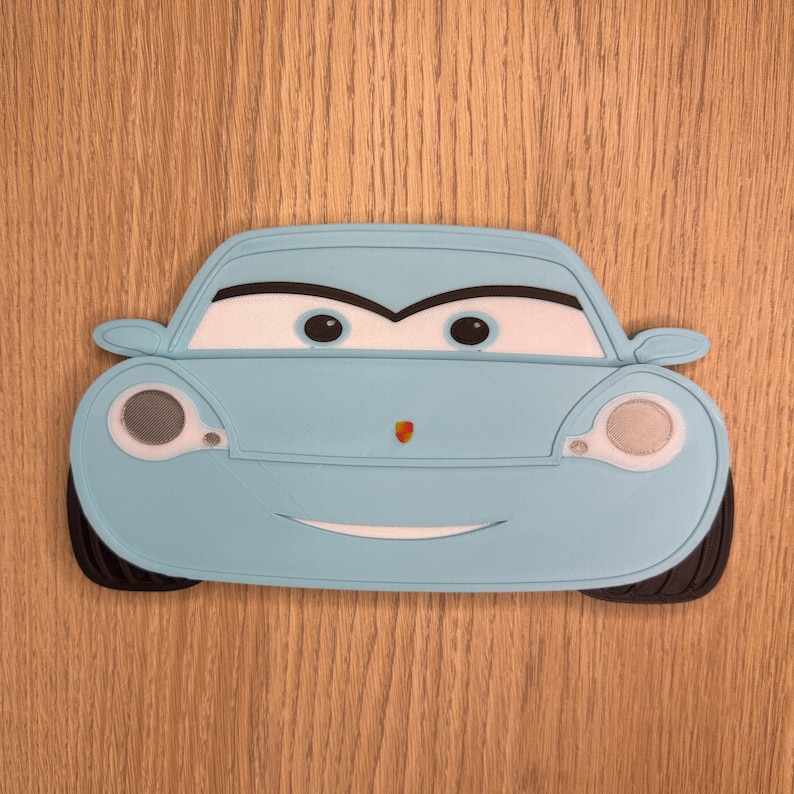 May include: A light blue cartoon car with a smiling face, black tires, and white headlights. The car has a red emblem on the front and is set against a wooden background. The car is a fun, playful design.