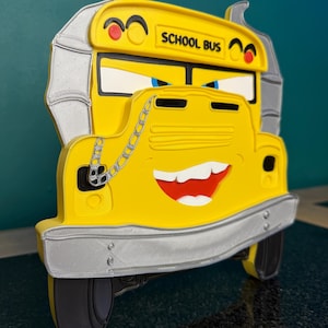 May include: A yellow school bus wall decoration featuring a cartoonish face with blue eyes, a wide grin, and silver exhaust pipes on the sides. A silver chain hangs over the hood, and the text SCHOOL BUS is printed in black on the front.