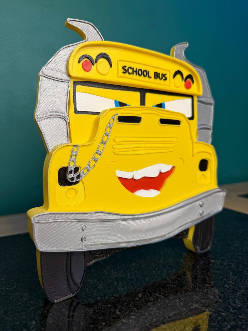 May include: A yellow school bus wall decoration featuring a cartoonish face with blue eyes, a wide grin, and silver exhaust pipes on the sides. A silver chain hangs over the hood, and the text SCHOOL BUS is printed in black on the front.