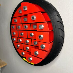 May include: A red and black advent calendar shaped like a tire. The calendar has 24 numbered compartments with silver-colored bolt-shaped knobs. The tire has a black rubber tread.