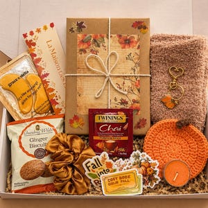 May include: A gift box containing a wrapped book with autumn leaf motifs, a pair of fuzzy socks, a gold heart keychain with a leaf charm, a crochet pumpkin coaster, a candle, ginger biscuits, a scrunchie, tea, eye masks, and stickers.