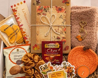 May include: A gift box containing a wrapped book with autumn leaf motifs, a pair of fuzzy socks, a gold heart keychain with a leaf charm, a crochet pumpkin coaster, a candle, ginger biscuits, a scrunchie, tea, eye masks, and stickers.