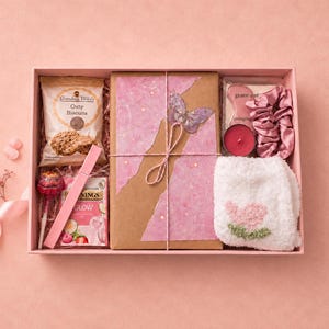 May include: A pink gift box containing a packet of Grandma Wild's Oaty Biscuits, a Twinings Glow tea box, a pink lollipop, a wrapped book with a butterfly cutout, a pink gua sha tool, a red candle, a pink scrunchie, and white fluffy socks.
