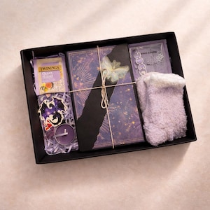 Könnte beinhalten: Eine schwarze Geschenkbox mit Artikeln zur Selbstpflege. Enthält ein Buch, Tee, Socken, eine Kerze und einen Aufkleber. Das Buch hat ein lila-goldenes Himmelsdesign. Auf der Teepackung steht "Twinings Quiet Mind".